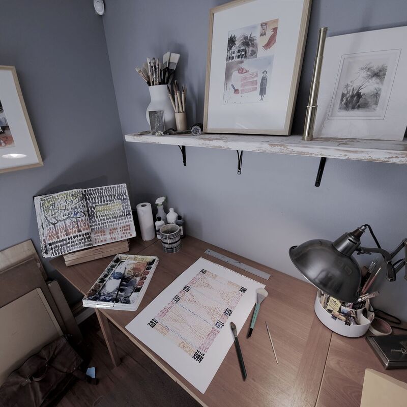 Photo of part of Bruce Black's art studio.  A lamp and various paintings are shown along with a watercolor palette. 